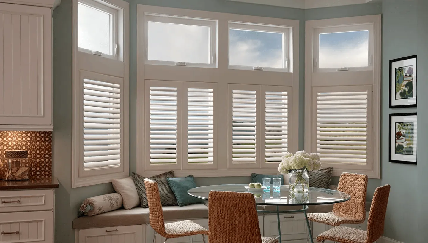 Kitchen breakfast nook bay windows fitted with white plantation shutters beneath transom windows around a round dining table.