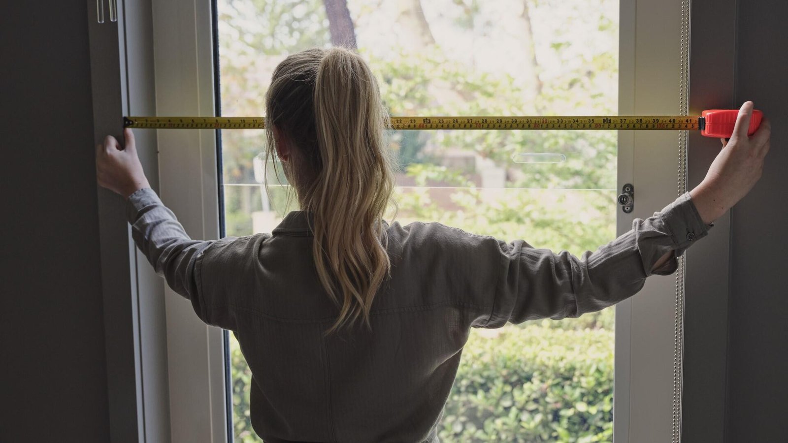 Woman using a yellow tape measure to measure the width of a glass window frame.